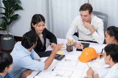 Mimari proje üzerinde birlikte çalışan çeşitli inşaat mühendisi ve müvekkil grupları, inşaat planını gözden geçirip toplantı masasında plan oluşturuyorlar. Tedbirli