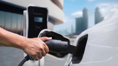 Adam elektrikli arabaya akıllı EV şarj cihazı takıyor ve arka planda modern şehir manzarası olan EV şarj istasyonundan şarj oluyor. ESG şehrinde gelecekteki sürdürülebilir güç ve EV araba kullanımı. İnceleyin