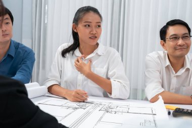 Mimari proje üzerinde birlikte çalışan çeşitli inşaat mühendisi ve müvekkil grupları, inşaat planını gözden geçirip toplantı masasında plan oluşturuyorlar. Tedbirli