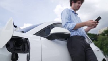 Takım elbiseli, hırslı bir işadamı elektrik kablosu ve yenilenebilir enerji enerjili bir elektrikli araçla şarj istasyonunda dururken elektrikli aracına yaslanıyor..