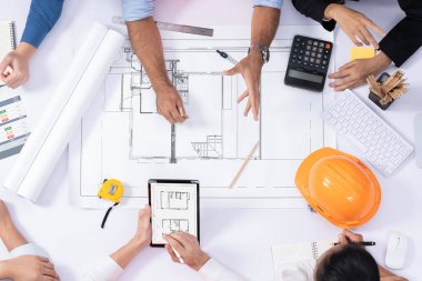 Mimari proje üzerinde birlikte çalışan, inşaat planını gözden geçiren ve toplantı masasında plan oluşturan çeşitli inşaat mühendisi ve müşteri gruplarından oluşan üst düzey bir pankart. Tedbirli