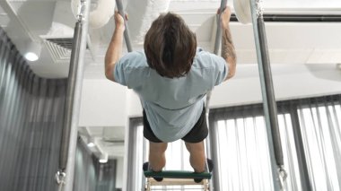 Man gripping bar on pilates trapeze table for core strength and upper body workout in a gym. Trainer hanging inverted back bend on the cadillac for improving flexibility in back and hip. Habituate. clipart