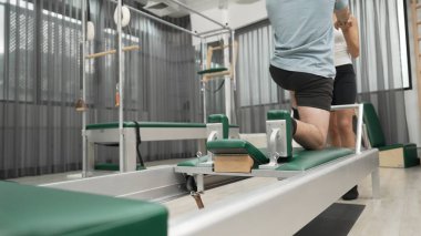 Esneklik ve dayanıklılığı artırmak için pilates reformcusu üzerinde diz çökme alıştırması yapan bir adam. Antrenör asistanı dengeyi arttırmak için yenilikçiye hamle yapıyor. Alışkanlık.