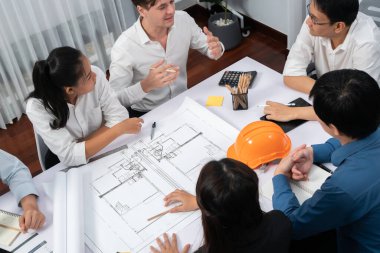 Mimari proje üzerinde birlikte çalışan çeşitli inşaat mühendisi ve müvekkil grupları, inşaat planını gözden geçirip toplantı masasında plan oluşturuyorlar. Tedbirli