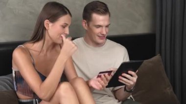 Genç çift, kredi kartıyla ödeme yapabilmek için internet üzerinden ödeme uygulaması ve dijital cüzdan kullanarak yatak odasında oturuyor. Mobil internet üzerinden alışveriş ve modern alımlar. Düzenle
