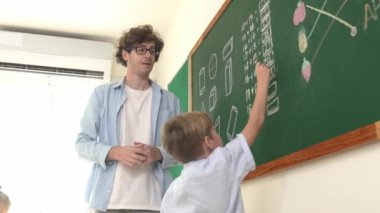 Beyaz çocuk tahtada cevap yazıyor ve sınıfın önünde dikilirken beşlik çakıyor. Sorulara cevap vermek için gönüllü olan zeki ilkokul öğrencilerini kapatın. Eğitim. Pedagoji.