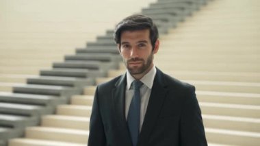 Akıllı iş adamı merdivenlerde kameraya bakıyor. İş elbisesi giyerken kameraya gülümseyen başarılı adamın yakın çekimi. Yönetici, kameraya bak. Hareket eden kamera. Sevinçli..