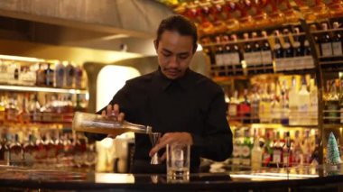 İyi görünümlü Asyalı barmenin içki şurubunu barın lüks içki tezgahında buzlu cama dönüştürdüğü profili. Gece kulübü lüksünde profesyonel barmen kostümü. Vinosity.