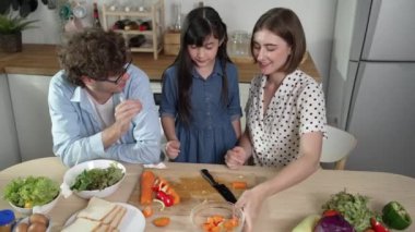 Çekici beyaz anne ve baba Asyalı kızıyla taze yemek pişirmek için birlikte vakit geçiriyorlar. Enerjik anne ve baba mutfakta salata yapmak için taze sebze hazırlıyor. Sağlıklı yiyecekler. Pedagoji.