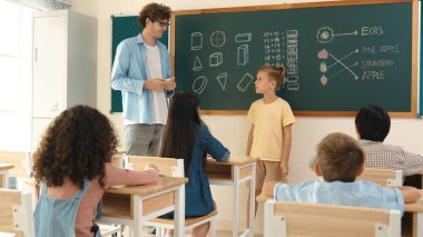 Profesyonel öğretmen konuşuyor ve sınıfta Asyalı çocuğa fikrini açıklıyor. Sınıfın önünde karatahta matematik dersiyle dikilmiş beyaz öğrenci dinleme eğitmeni. Pedagoji.
