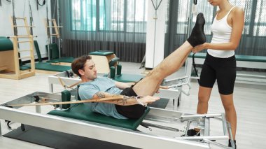 Bir grup müşteri, yenilikçi bir makinede bacak yükseltme işlemi yapıyor. Pilates partneri reformcu pozu veriyor ve egzersiz sırasında dengeyi geliştirmek ve koordinasyonu arttırmak için kayış takıyor. Alışkanlık.