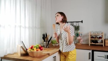 Enerjik beyaz anne kahvaltı için salata pişirirken ve yemek pişirirken elinde kepçeyle şarkı söylüyor. Sağlıklı yemek yapmaya hazırlanırken müziğe yönelen mutlu ev sahibi. Pedagoji.