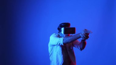 Meta-World 'de atış oyunu oynarken VR kulaklık takan profesyonel bir oyuncu. Erkek model, sanal gerçeklik gözlüğü kullanarak mavi arka planda dururken silah hareketi yapıyor. Aygıt.