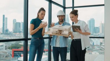 Proje yöneticisi ev modelini elinde tutarken, mimar mühendis takımı da ev tasarımı veya bina inşaatı hakkında fikir alışverişinde bulunuyor. Şehrin penceresinin yanında iç mimar duruyor. İzleme