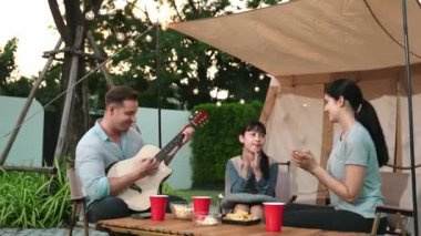 Baba gitar çalarken anne ve kız birlikte kampta oturuyorlar. Ebeveynler, genç nesil çapraz kuşak farklılıklarıyla zaman geçirmek için açık hava kamp aktivitelerini kullanıyorlar. Uyumsuzluk.