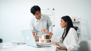 Profesyonel Asyalı erkek mimar ev modelinin uzunluğunu ölçmek için cetvel kullanırken, beyaz tenli genç meslektaşı ev modeliyle toplantı masasındaki verileri analiz etmek için dizüstü bilgisayar kullanıyor. Tertemiz..