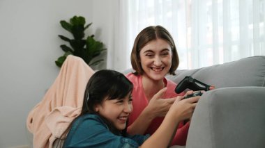 Annem dizüstü bilgisayar kullanarak kanepeye uzanırken, mutlu kız oyun oynuyor ve oturuyor. Beyaz bir ebeveyn ve çekici bir kız, elinde joystick ile birlikte vakit geçirir ve maç kazanmaya odaklanırlar. Pedagoji.