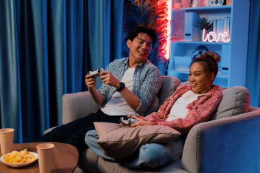 Mutlu çiftin oturduğu kanepe joysticklerle video oyunu oynuyor, meydan okuyan gülümseyen adam, evdeki modern neon ışıklı oturma odasında kazanmak için yumruğunu kaldırıyor. İnfobahn.