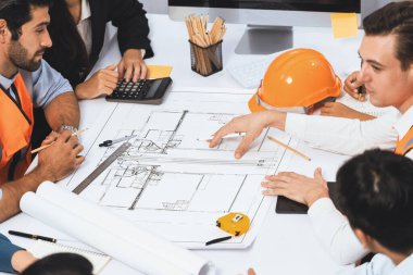 Mimari proje üzerinde birlikte çalışan çeşitli inşaat mühendisi ve müvekkil grupları, inşaat planını gözden geçirip toplantı masasında plan oluşturuyorlar. Tedbirli