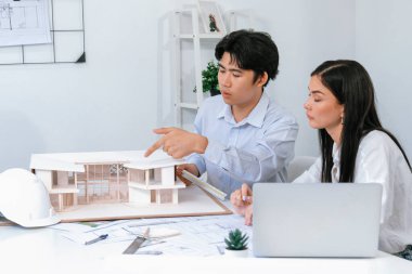Profesyonel Asyalı erkek mimar ev modelinin uzunluğunu ölçmek için cetvel kullanırken, beyaz tenli genç meslektaşı ev modeliyle toplantı masasındaki verileri analiz etmek için dizüstü bilgisayar kullanıyor. Tertemiz..