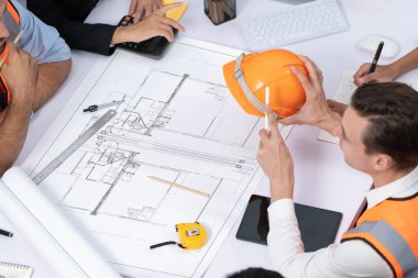 Mimari proje üzerinde birlikte çalışan çeşitli inşaat mühendisi ve müvekkil grupları, inşaat planını gözden geçirip toplantı masasında plan oluşturuyorlar. Tedbirli