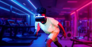 Afrikalı bir adam parlak renkli fütüristik spor salonunda spor yaparken VR gözlük takıyor ve rahat bir gömlek giyiyor. Koşu pozu veren erkek model. Teknoloji yeniliği. Sağlıklı bir yaşam tarzı. Yaratıcılık.