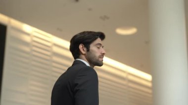 Profesyonel bir iş adamı kolları bağlıyken arkasını döner. Kafkasyalı mutlu yatırımcı, arka planı bulanık bir şekilde koridorda kollarını kavuştururken kameraya özgüvenle bakıyor. Portre. Sevinçli..