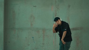Enerjik hipster şehir caddesinde kulaklıktan hip-hop müziğiyle dans ediyor. Sokak dansı performansı çalışırken kameraya bakan break dansçı. Geçmişi. Açık hava sporu 2024. Hiphop.