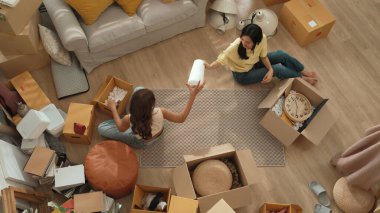 Oturma odasındaki mobilyaların taşınmasına yardım ederken yerde oturan mutlu oda arkadaşı. Yeni eve taşınmak için birlikte çalışan Asyalı lezbiyen aşığının havadan görünüşü. Yer değiştirme konsepti. Myrmidon.