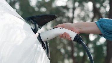 Kapalı alan kadını, doğal yeşil parktaki otoparkta EV elektrikli araba aküsünü şarj ediyor. Sürdürülebilir ve çevre dostu seyahat için şarj edilebilir araç için temiz enerji teknolojisi. Titiz