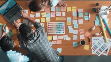 Gündelik kıyafet giyen, dizüstü bilgisayara bakan ve fikir paylaşan iş ekibinin zaman atlaması. Yapışkan notlar ve kulaklıkla masada çalışan ve renk paletini kontrol eden yöneticinin hava görüntüsü. Sempozyum.