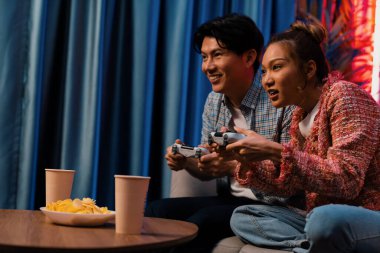 Neşeli, gülümseyen bir çiftin koltuğunda oturup oyun oynuyorlar. Oyun konsolu, hazır aperatifler ve içecekler, modern neon ışıklı, rahat oturma odasında eğlenceli bir rekabet seviyesinde. İnfobahn.