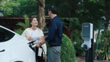 Yavaş çekim çift, ilkbaharda açık yeşil şehir kafesindeki şarj istasyonundan elektrikli araba aküsünü şarj ediyor. Sürdürülebilir çevre dostu seyahat yaşam tarzı için şarj edilebilir EV araba