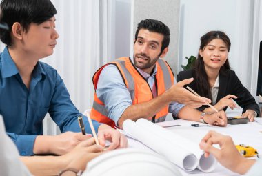 Mimari proje üzerinde birlikte çalışan çeşitli inşaat mühendisi ve müvekkil grupları, inşaat planını gözden geçirip toplantı masasında plan oluşturuyorlar. Tedbirli