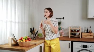 Enerjik beyaz anne kahvaltı için salata pişirirken ve yemek pişirirken elinde kepçeyle şarkı söylüyor. Sağlıklı yemek yapmaya hazırlanırken müziğe yönelen mutlu ev sahibi. Pedagoji.