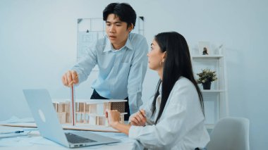 Professional male asian architect using ruler to measure house model length while young beautiful caucasian colleague using laptop to analyzed data on meeting table with house model. Immaculate. clipart