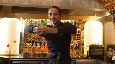 Yakın plan Asyalı barmen, lüks tezgahta karışık kokteyl sallayarak kamera önüne bakarak özel içecek menüsü hazırlıyor. Gece hayatını gösteren özel bir içecek. Vinosity.