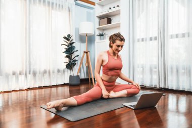 Spor kıyafetleri içinde esnek ve becerikli bir kadın evde egzersiz paspası üzerinde meditasyon pozisyonunda yoga yapıyor. Huzurlu bir zihin ve sükunetle evde yoga eğitimi..