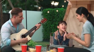 Baba gitar çalarken anne ve kız birlikte kampta oturuyorlar. Ebeveynler, genç nesil çapraz kuşak farklılıklarıyla zaman geçirmek için açık hava kamp aktivitelerini kullanıyorlar. Uyumsuzluk.