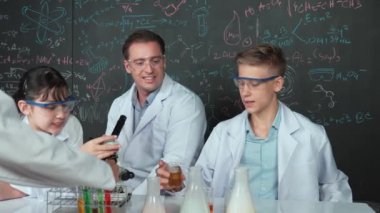 Laboratuvar önlüğü giyen genç bir çocuk, STEM fen sınıfında tahtada renkli sıvıyla bekletiliyor. Öğretmen masada tavsiye verirken bir grup farklı öğrenci deney yapıyor. Öğretim