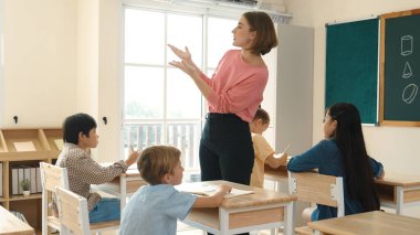 Beyazca öğretmeni, ilkokulda karatahta başında dururken vücut jesti kullanarak çok kültürlü öğrencilere teoriyi öğretiyor ve açıklıyor. Yetenekli öğretmen çocuklara soru soruyor. Pedagoji.