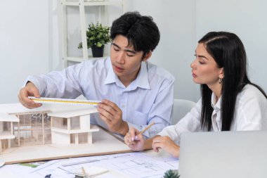 Profesyonel Asyalı erkek mimar ev modelinin uzunluğunu ölçmek için cetvel kullanırken, beyaz tenli genç meslektaşı ev modeliyle toplantı masasındaki verileri analiz etmek için dizüstü bilgisayar kullanıyor. Tertemiz..