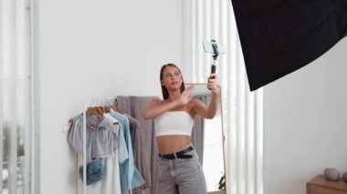 Genç sosyal medyum kadın moda videosu yapıyor. Blogcu kameraya selfie çubuğu ve ışık halkası kullanarak gülümserken, aynı zamanda ikna edici çevrimiçi kıyafetlerin izleyicilere veya takipçilere vlog satmasını sağlıyor. Düzenle
