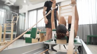 Eğitmen, müşteriye pilates reformatöründe rehberlik ediyor. Vücut geliştirme egzersizinde, vücut hareketlerine, esnekliğine ve beden kuvvetine odaklanmak için kayışlar kullanıyor. Asyalı kız pilates makinesinin önünde yatıyor. Alışkanlık.
