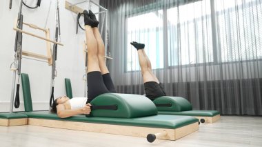 Pilates seansı sırasında iki müşteri yay namlu teçhizatı kullanarak bacak kaldırma pratiği yapıyor. Mutlu antrenör grubu karın kaslarını ve alt karın kaslarını geliştirmek için makas ya da bacak esneme pozu verir. Alışkanlık.