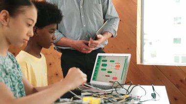 Öğretmen ayakta dururken kodlamayı ve robot yapısını açıkla. Çeşitli gençler ana kartı tamir ediyor ve dizüstü bilgisayarın programlama ekranına bakarken elektronik alet kullanıyorlar. Öğretim