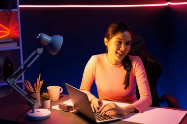 Geceleri sosyal medyada canlı yayında pozitif podcastla konuşan gülümseyen genç, güzel Asyalı etkileyici pembe neon ışıklı modern stüdyoda sunum için dizüstü bilgisayar kullanıyor. Taktik.