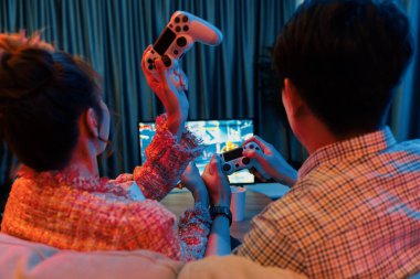 Kontrollü joystickli birkaç oyuncu arka tarafta televizyon ekranında dövüş oyunu oynuyor, modern rahat evinde neon ışıklı renkli oturma odasında koltukta oturuyor. İnfobahn.