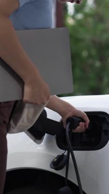 Kadın elektrikli arabasını akıllı ev şarj istasyonunda bulunan EV şarj cihazıyla şarj ediyor. Fütürist dijital batarya durumu hologramı. Sürdürülebilir güç için EV araba ve ev enerjisi altyapısı. İnceleyin