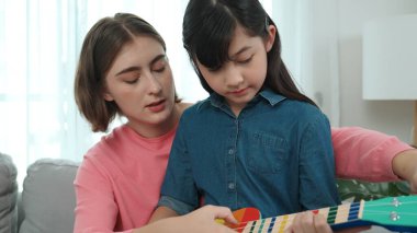 Kafkasyalı bir anne evde akustik müzik öğretirken ukulele çalan mutlu kız. Sevimli çocuk enstrüman öğreniyor. Kafkasyalı mutlu anne ve kız birlikte vakit geçiriyorlar. Pedagoji.